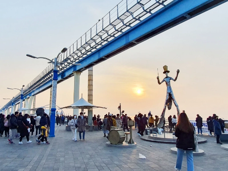 夕焼けに染まる月尾文化通りの海沿いの遊歩道と、行き交う人でにぎわう港の風景